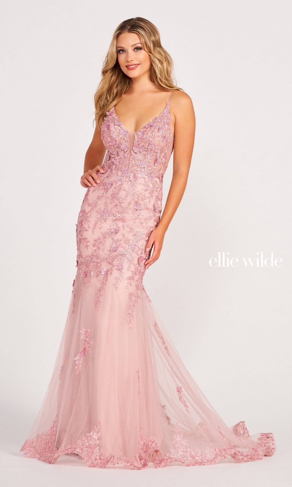 A woman wears the Lace Embroidered Mermaid Gown With Sheer Bodice, a light pink, floor-length lace dress with floral embroidery, sheer corset details, and thin straps, posing against a plain white background.